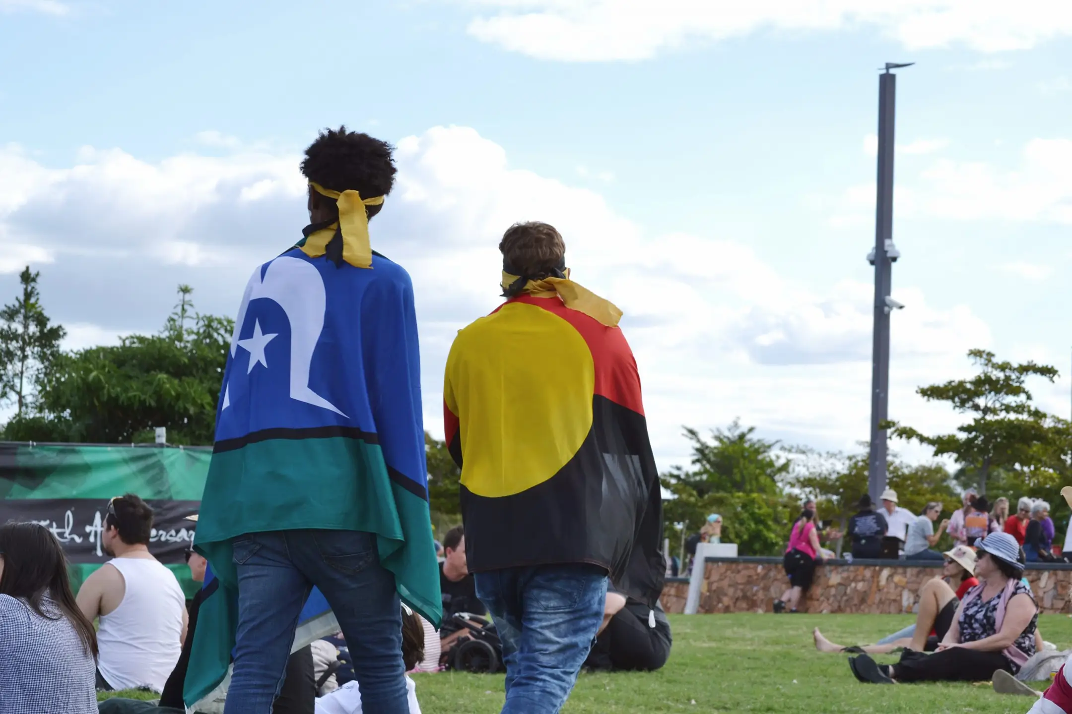 Two individuals are seen from the back, draped in flags representing different cultural heritages, walking through a park where various other people are sitting and enjoying the day. The person on the left is wearing a Torres Strait Islander flag, while the person on the right is wrapped in an Aboriginal flag. The scene is casual and appears to be during a public event or gathering