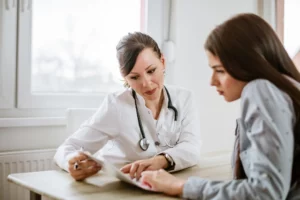 A doctor seated at a table across from a patient, deeply engaged in a conversation. The doctor, wearing a white coat and a stethoscope, is focused on a document on the table, possibly discussing the patient's health information or test results. The patient appears attentive and concerned, listening closely to the doctor's explanations. The setting suggests a private consultation room with natural light coming in from a window.